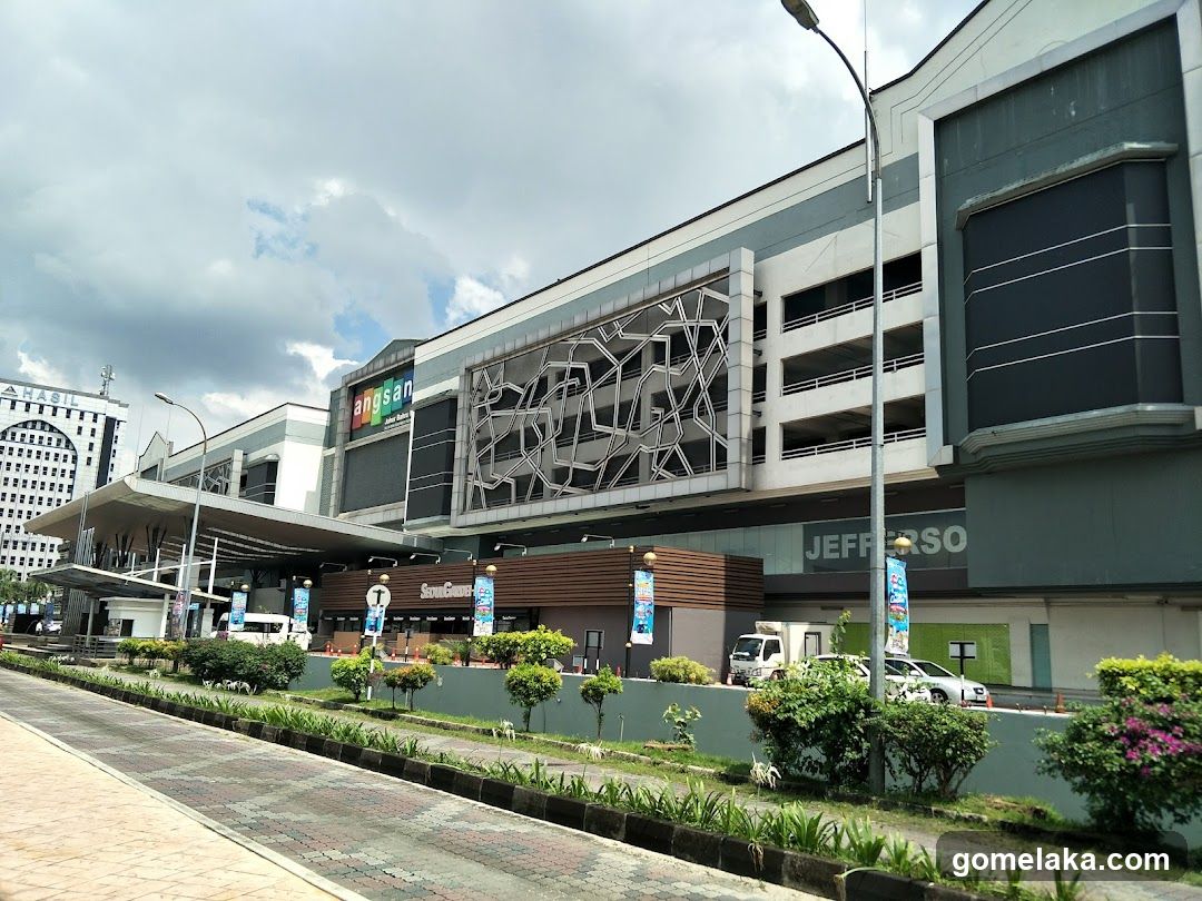 A wide shot of the exterior of a large shopping mall. The mall has a modern design with a grey and white facade. A large sign with colorful letters that reads "angsan" is visible at the top of the building. Below this, there is a section with a geometric pattern. Further to the right, the word "JEFFERSO" is visible in large letters on the building. The entrance area has a covered walkway and several trees and bushes line the front. A white van and a silver car are parked in front of the mall.