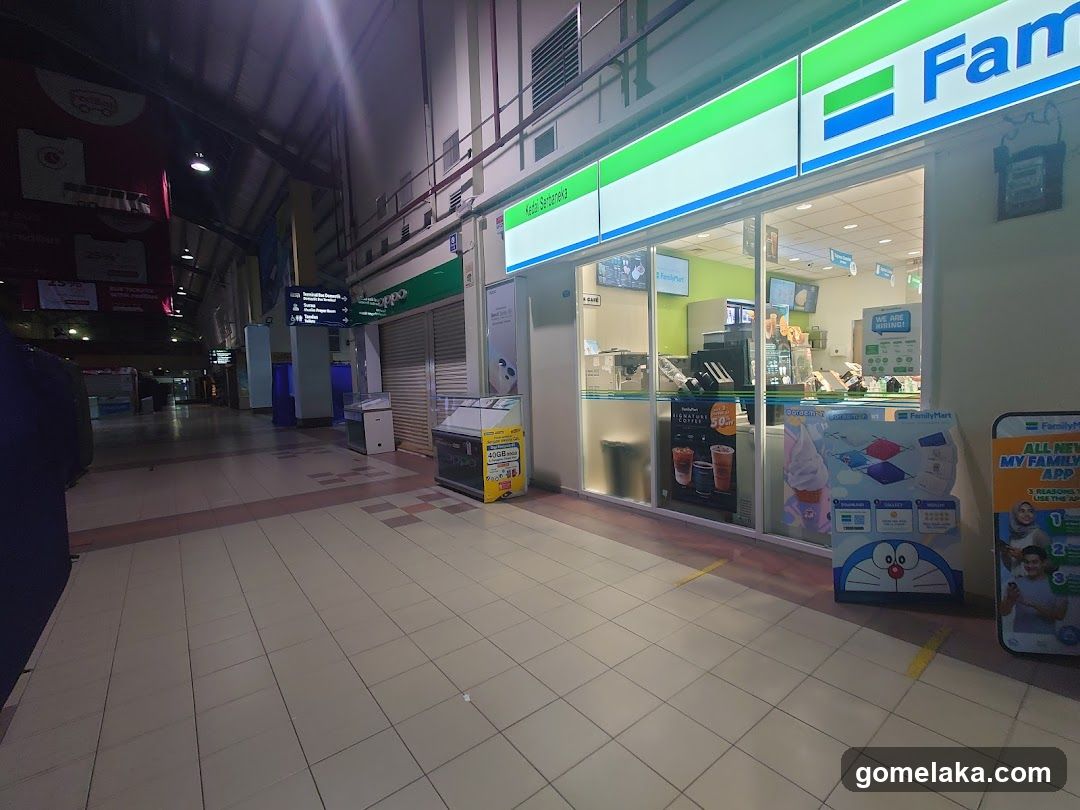 A brightly lit FamilyMart store with green and blue signage above the entrance. The storefront has large glass windows showing the interior, including a counter, coffee machines, and promotional displays. A Doraemon-themed standee is placed outside near the entrance. Adjacent to it, another standee promotes the 'My Family App'. To the left, a shuttered shop with a green sign is visible. The floor is tiled, and overhead lighting illuminates the area.