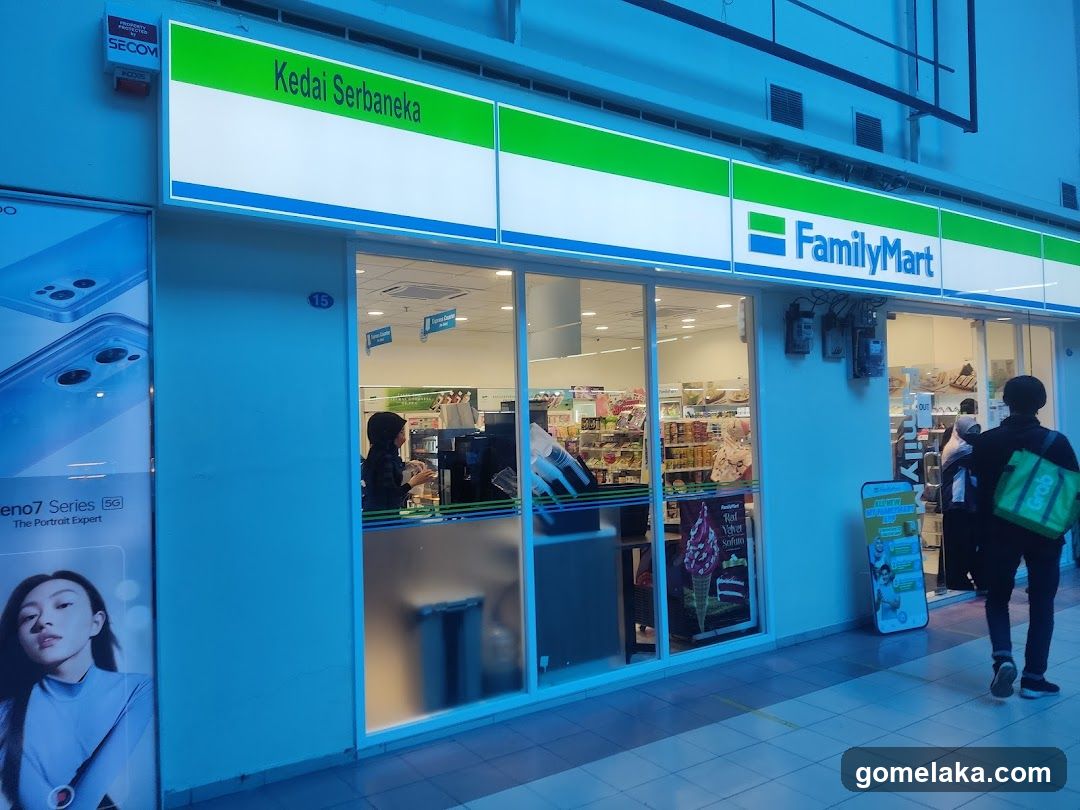 Exterior view of a FamilyMart store with green and white signage above the entrance. Large glass windows display interior shelves and products. A customer wearing a green bag walks past the entrance. The building facade is painted light blue, with a SECOM alarm box mounted on the wall.