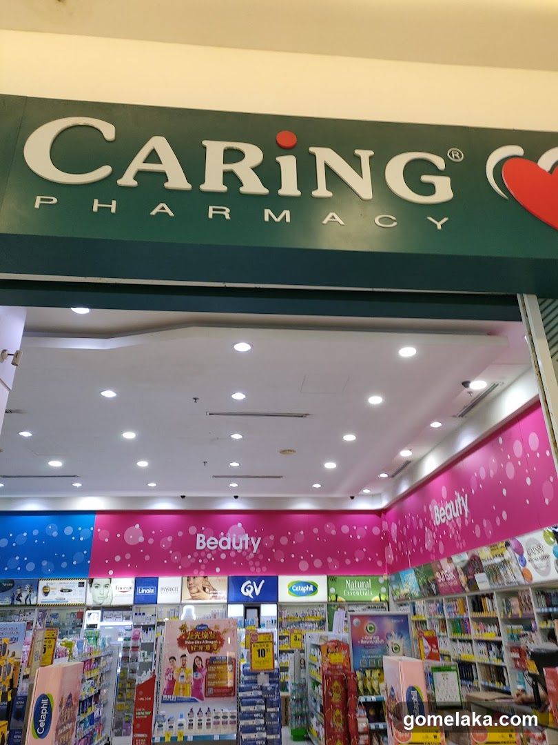 Storefront with green sign featuring 'CARING' in white letters and 'PHARMACY' below it; red heart icon to the right. View from outside looking into brightly lit interior with pink 'Beauty' section signage, shelves stocked with products, and brand logos like QV, Cetaphil, and Linolei visible inside.