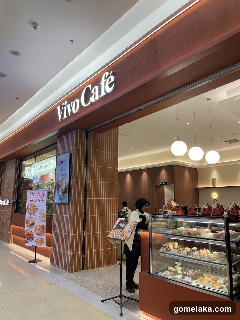 Modern interior-facing entrance with illuminated 'Vivo Café' sign above the counter. Glass display case showcases pastries and cakes. Staff member stands beside a promotional stand holding a printed menu or catalog. The shop features warm lighting, wooden paneling, and hanging spherical lights inside.