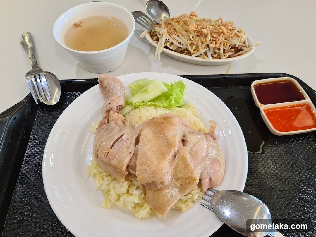 A meal served on a black tray, featuring Hainanese chicken rice with sliced chicken, rice, cucumber slices, and lettuce. Accompanied by a bowl of clear broth, a side dish of bean sprouts topped with fried shallots, and two small sauce containers (dark red and orange-red). Silverware is placed around the plate.