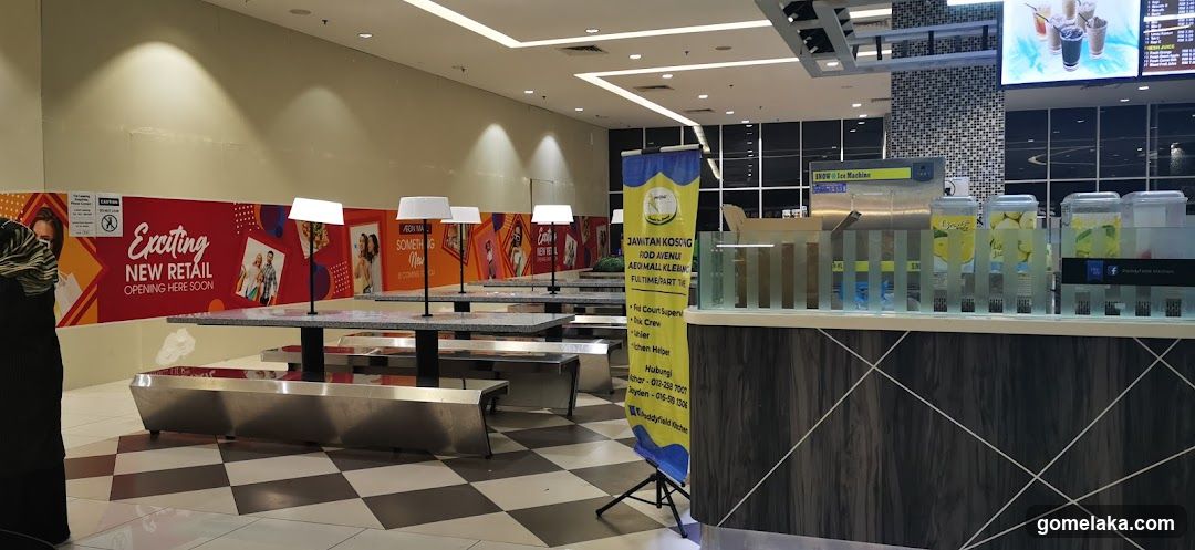 Interior of a shopping mall food area with checkered flooring, metal tables, and a beverage station featuring lemon juice dispensers. A person in hijab stands near the left. Brightly colored retail banners advertise 'Exciting NEW RETAIL OPENING HERE SOON'. A yellow banner lists contact info for 'JAWATAN KOSSIG' (likely local service). Large digital screens show drink images above.