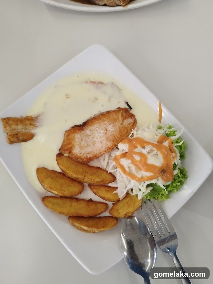 A square white plate with a meal consisting of fried fish fillet in creamy sauce, roasted potato wedges, and a side salad with shredded cabbage, lettuce, and orange drizzle. A spoon and fork are placed beside the plate on a light-colored surface.