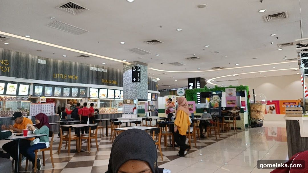 Interior of a food court with multiple dining tables, chairs, and patrons. Visible signage includes 'LITTLE WOK' and 'THAI & GRILL'. A few people are seated or walking. Background features a green wall display and promotional posters.