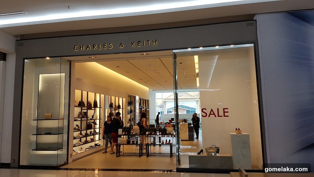 Modern glass storefront with illuminated 'CHARLES & KEITH' signage above entrance. Interior visible through glass, showing shelves of shoes and bags, sale sign on right wall, customers browsing inside.
