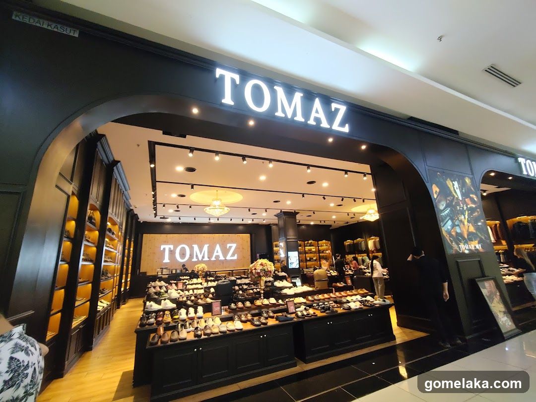Modern retail entrance with illuminated 'TOMAZ' sign above arched entryway. Interior features black display counters, wooden shelving, and bright lighting. Customers are visible inside the store.