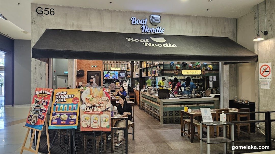 Storefront with concrete facade, black awning, illuminated 'Boat Noodle' sign above entrance. Interior visible with staff at counter, customers seated, brick wall background, no external view.