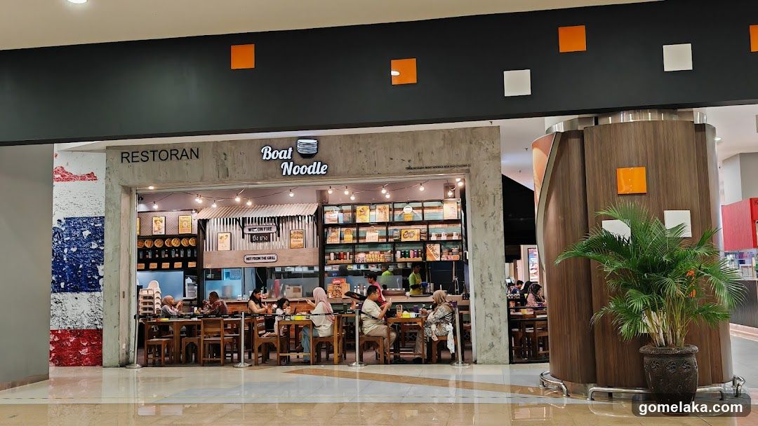 A storefront with a concrete facade and black ceiling adorned with orange and white square accents. The shop name "Boat Noodle" is displayed in illuminated lettering above the entrance. Inside, patrons are seated at wooden tables under string lights, with shelves displaying products behind the counter.