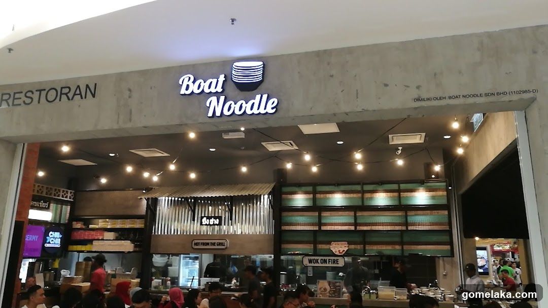 The storefront features a concrete facade with illuminated "Boat Noodle" signage above the entrance. The sign includes a stylized noodle bowl logo. To the left, partial text reads "RESTORAN". Below, string lights hang from the ceiling inside the restaurant, and a "WOK ON FIRE" sign is visible in the kitchen area. Customers are seated at tables inside, and staff are present behind counters.