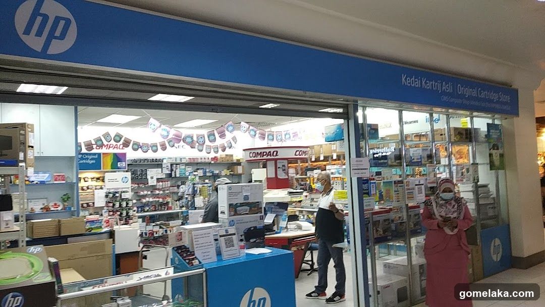Storefront with blue HP signage, glass doors, and visible interior shelves stocked with products. A person in a face mask stands near the entrance. Signage reads 'Kedai Kartriji Asli - Original Cartridge Store'.