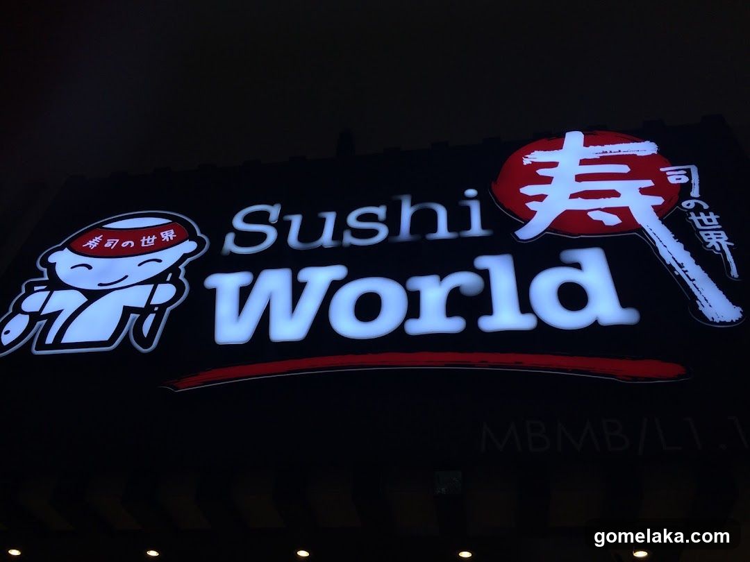 Nighttime photo of a brightly lit illuminated sign on a dark building facade. The sign features the name 'Sushi World' in large white letters, accompanied by a cartoon sushi chef mascot wearing a red hat with Japanese characters and a large red Japanese character (寿) next to it. Below the main logo is a red underline. Partially visible text at bottom right reads 'MBMB/L1'.