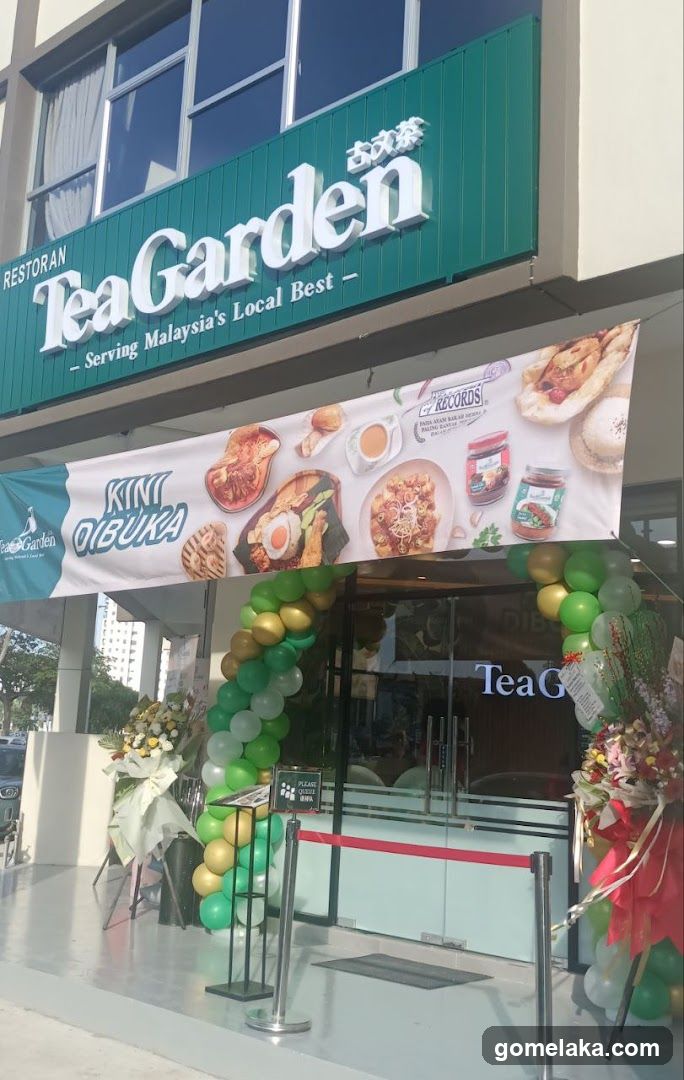 Exterior storefront with green signage reading 'Tea Garden - Serving Malaysia's Local Best'. A large banner displays food items and 'KINTO IBUKA' branding. Green and gold balloons decorate the entrance, flanked by floral arrangements. Glass doors have partial 'TeaG' visible.
