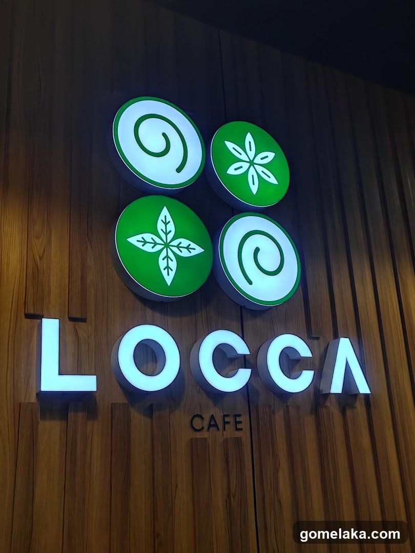 Illuminated logo on a wooden panel wall featuring four green circular icons with white designs (two spirals, two leaf-like patterns) above the glowing 'LOCCA' and 'CAFE' text.