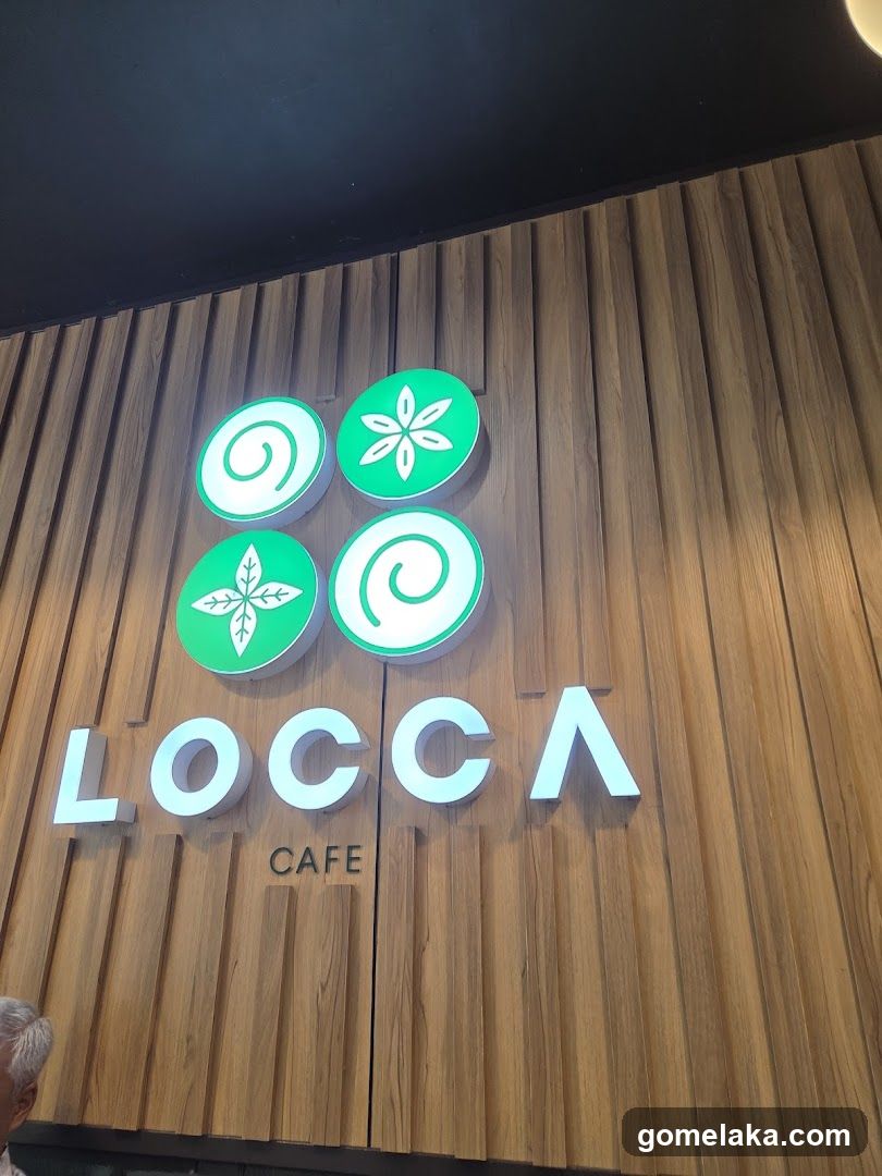 A modern, illuminated sign on a wooden panel wall featuring the name 'LOCCA' and 'CAFE' below it. Above the name are four green circular logos with white symbols: two spirals and two leaf-like designs. The background is a vertical wood-grain facade under a dark ceiling.